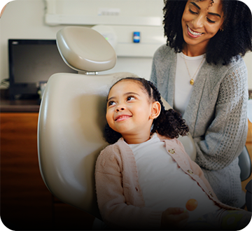 Little girl in dental chair with mother standing next to her